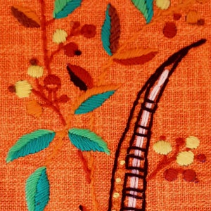 May include: Close-up of embroidered floral design on orange fabric. The embroidery features teal and green leaves, yellow flower clusters, and a black and white striped element. The design is detailed and colorful.