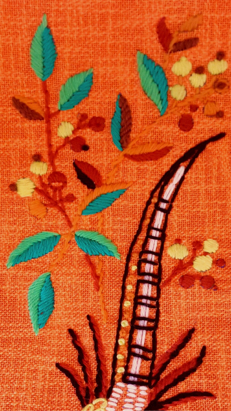 May include: Close-up of embroidered floral design on orange fabric. The embroidery features teal and green leaves, yellow flower clusters, and a black and white striped element. The design is detailed and colorful.