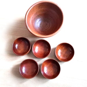 May include: A set of six wooden bowls. One large bowl is positioned above five smaller bowls. The bowls are made of a reddish-brown wood and have a smooth, polished finish. The bowls are empty and arranged on a light-colored wooden surface.