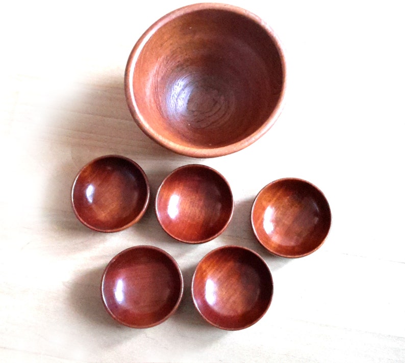 May include: A set of six wooden bowls. One large bowl is positioned above five smaller bowls. The bowls are made of a reddish-brown wood and have a smooth, polished finish. The bowls are empty and arranged on a light-colored wooden surface.