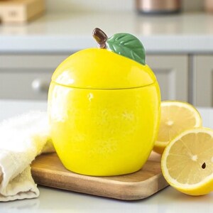 May include: A vibrant yellow ceramic lemon-shaped jar, featuring a green leaf and brown stem. It rests on a wooden board, accompanied by two lemon halves. The jar has a shiny, glazed surface.