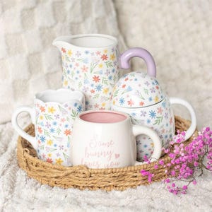 May include: A set of floral-patterned ceramic mugs, a creamer jug, and a lidded mug, presented on a woven tray. The white mugs and jug feature colourful flower designs. One mug has the text "Some bunny loves you" in pink.