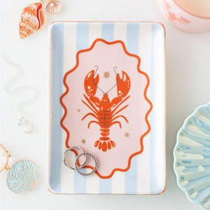 May include: Rectangular ceramic trinket tray with a light blue and white striped background. The tray features a pink oval with an orange lobster illustration. Two silver rings rest on the tray. Gold trim edges the tray.