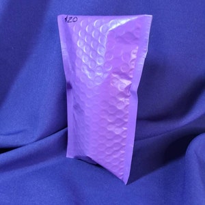 Puede incluir: Un peque&ntilde;o sobre de burbujas morado con una superficie texturizada. El sobre est&aacute; sellado y parece contener un art&iacute;culo. El fondo es una tela azul oscuro lisa.