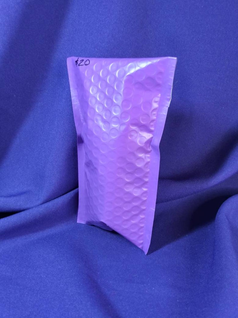 Puede incluir: Un peque&ntilde;o sobre de burbujas morado con una superficie texturizada. El sobre est&aacute; sellado y parece contener un art&iacute;culo. El fondo es una tela azul oscuro lisa.