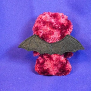 May include: A plush red toy with black felt bat wings. The toy is made of a soft, fuzzy material.