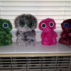 Puede incluir: Cuatro peluches en fila, cada uno con ojos grandes y redondos. Los juguetes son verdes, grises, rosas y burdeos. Los ojos tienen detalles brillantes. Los juguetes tienen un aspecto difuso y texturizado.