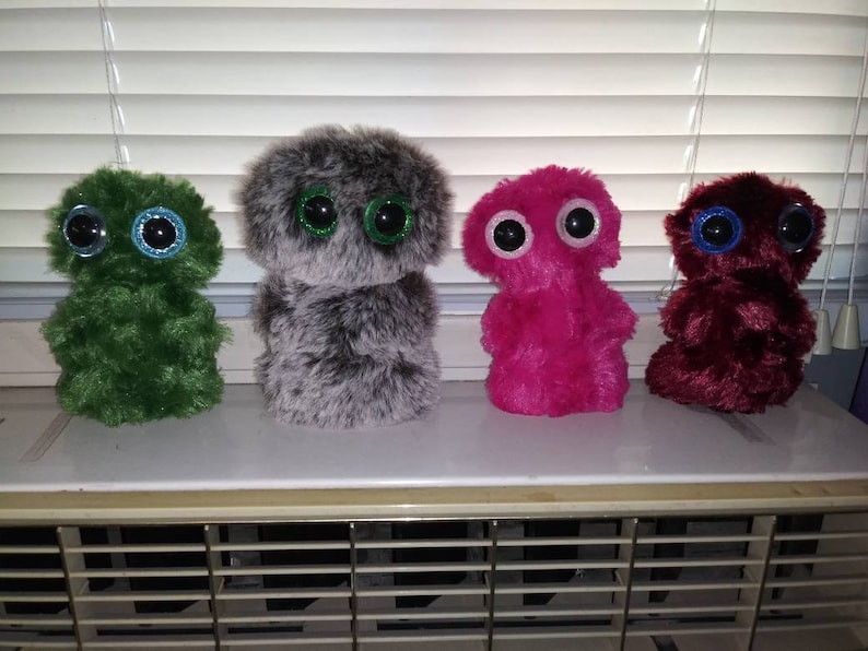 Puede incluir: Cuatro peluches en fila, cada uno con ojos grandes y redondos. Los juguetes son verdes, grises, rosas y burdeos. Los ojos tienen detalles brillantes. Los juguetes tienen un aspecto difuso y texturizado.