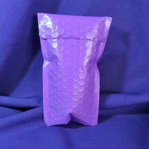 Puede incluir: Un sobre de burbujas morado se alza sobre un fondo azul oscuro. El sobre est&aacute; hecho de un material texturizado con un patr&oacute;n de peque&ntilde;os c&iacute;rculos en relieve. La parte superior est&aacute; doblada y la inferior sellada.