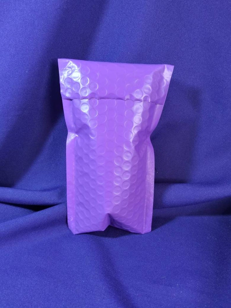 Puede incluir: Un sobre de burbujas morado se alza sobre un fondo azul oscuro. El sobre est&aacute; hecho de un material texturizado con un patr&oacute;n de peque&ntilde;os c&iacute;rculos en relieve. La parte superior est&aacute; doblada y la inferior sellada.