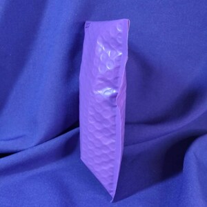 Puede incluir: Un sobre de burbujas morado, sellado y listo para enviar. La superficie tiene una textura. El fondo es una tela azul liso.