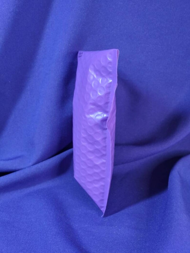 Puede incluir: Un sobre de burbujas morado, sellado y listo para enviar. La superficie tiene una textura. El fondo es una tela azul liso.