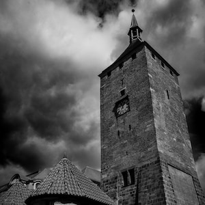 May include: Black and white photograph of a tall stone tower with a clock, part of a historic building. The tower has a pointed roof and is set against a dramatic, cloudy sky. A smaller building with a tiled roof is in the foreground.