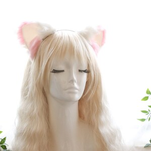 Puede incluir: Una diadema blanca con orejas de gato de pelo rosa. La diadema est&aacute; en un maniqu&iacute; con el pelo largo y rubio.