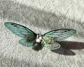 May include: A green and silver butterfly brooch with a clear rhinestone in the center.