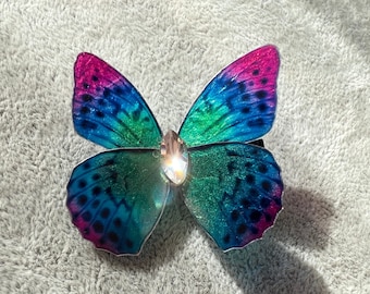 May include: A blue and green butterfly hair clip with a sparkly pink and purple iridescent finish. The clip has a clear rhinestone in the center.