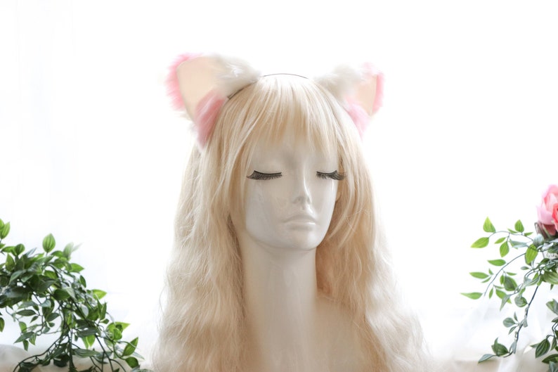 Puede incluir: Una diadema blanca con orejas de gato de pelo rosa. La diadema est&aacute; en un maniqu&iacute; con el pelo largo y rubio.