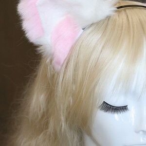 Puede incluir: Una diadema de orejas de gato de piel sint&eacute;tica blanca y rosa. La diadema se lleva en un maniqu&iacute; con el pelo largo y rubio.