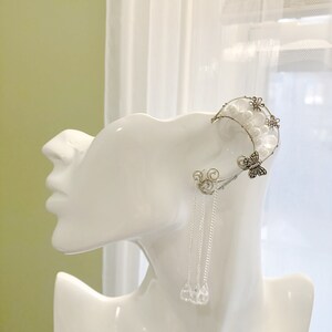Puede incluir: Un pendiente de oreja de plata con un diseño de media luna, adornado con perlas blancas y un dije de mariposa. El pendiente se combina con un arete colgante con una cadena de plata y tres gotas de cristal transparente.