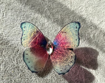 May include: A iridescent butterfly hair clip with a clear rhinestone in the center. The wings are a gradient of pink, blue, and green.
