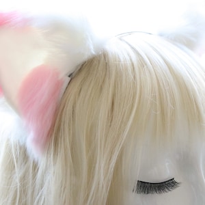 Puede incluir: Una diadema con orejas de gato de peluche blancas y rosas. La diadema est&aacute; puesta en un maniqu&iacute; con cabello rubio y ojos cerrados.