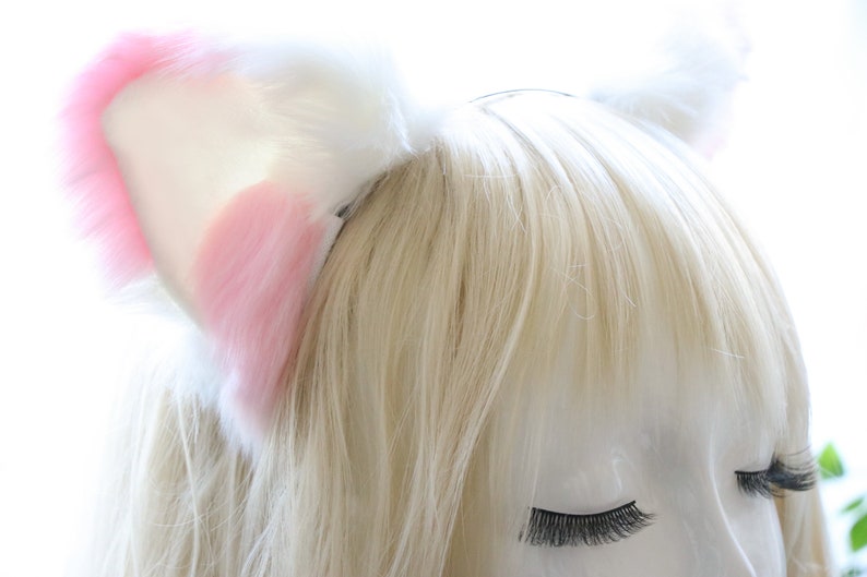 Puede incluir: Una diadema con orejas de gato de peluche blancas y rosas. La diadema est&aacute; puesta en un maniqu&iacute; con cabello rubio y ojos cerrados.