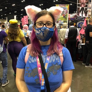 Puede incluir: Una persona con el pelo rosa que lleva una mascarilla facial azul y una camiseta azul con un perro de dibujos animados. Lleva una correa negra con un cord&oacute;n rosa y azul con el texto "Operaci&oacute;n: Amabilidad" en &eacute;l. Tambi&eacute;n lleva un par de orejas de gato blancas.