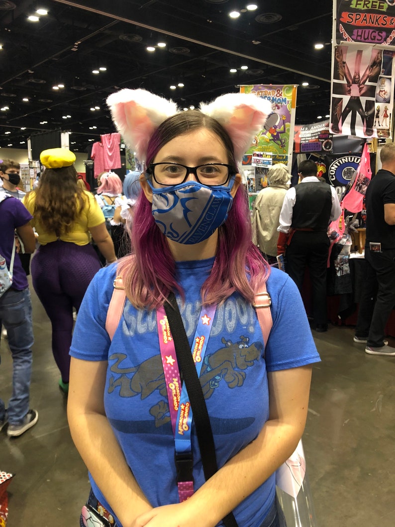 Puede incluir: Una persona con el pelo rosa que lleva una mascarilla facial azul y una camiseta azul con un perro de dibujos animados. Lleva una correa negra con un cord&oacute;n rosa y azul con el texto "Operaci&oacute;n: Amabilidad" en &eacute;l. Tambi&eacute;n lleva un par de orejas de gato blancas.