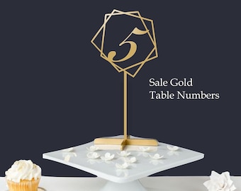 May include: Gold table number five in a geometric frame on a gold stand. The number is on a white cake stand with white flowers. The text "Sale Gold Table Numbers" is below the number.