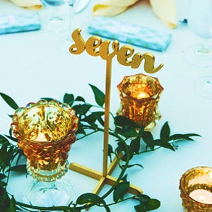 Può includere: Un numero di tavolo dorato con la parola "Seven" su di esso, circondato da verde e tre portacandele dorati con candele accese.