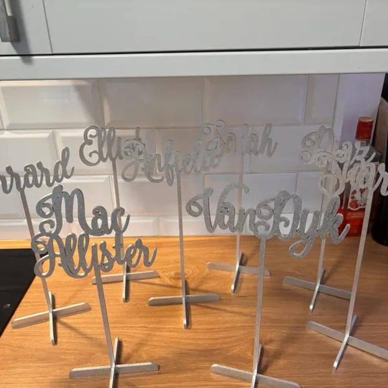 May include: Silver glitter table name cards with names including Gerard, Ellie, Alfred, Sarah, MacAllister, Van Dyk, and Blair. The cards are on silver stands.