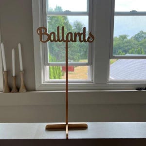 May include: A wooden table number sign with the word "Ballards" in gold lettering. The sign is on a wooden stand with a cross-shaped base.