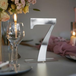 May include: A mirrored silver number seven table decoration. The number is displayed on a clear acrylic base, set on a white surface. The background includes a floral arrangement, candles, and glassware, suggesting a wedding or event setting.