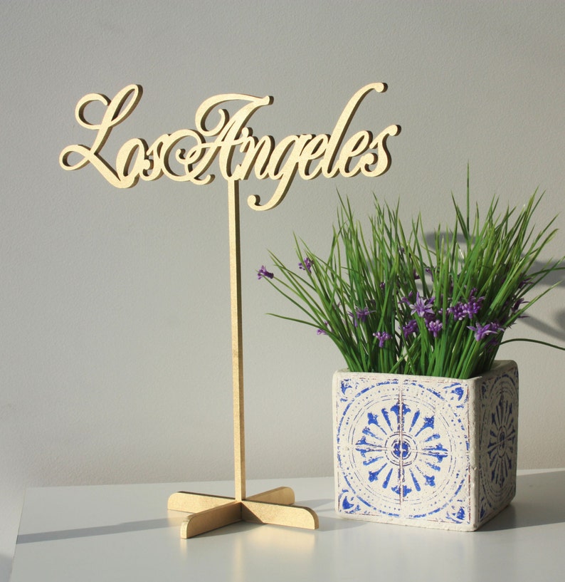 May include: A wooden sign with the words "Los Angeles" in cursive script, mounted on a stand. The sign is in front of a potted plant with purple flowers and a blue and white patterned planter.