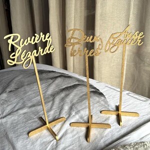 May include: Three gold table number signs with the words "Riviere Lezarde", "Deux Terres", and "Rose Baguier" written in cursive.