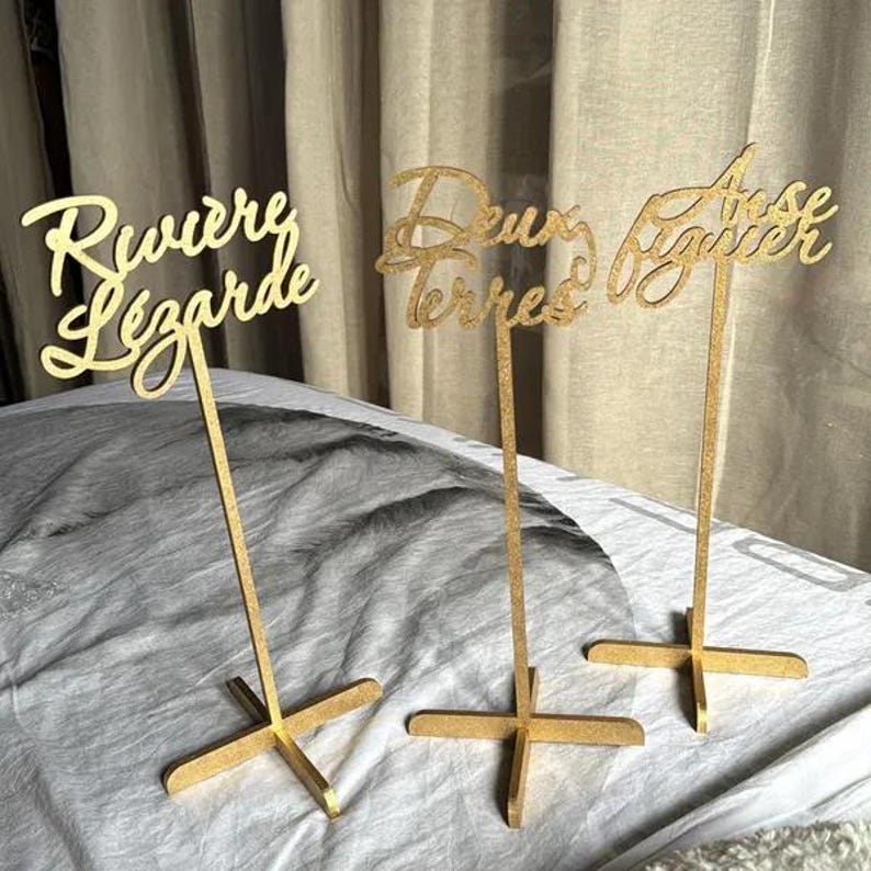 May include: Three gold table number signs with the words "Riviere Lezarde", "Deux Terres", and "Rose Baguier" written in cursive.