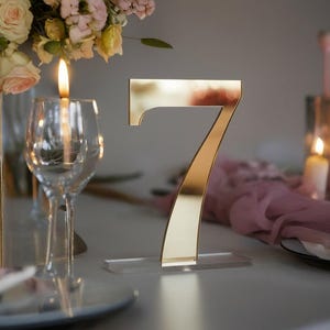 Può includere: Un numero da tavolo dorato, il numero sette, è posto su una base in acrilico trasparente. Il numero è esposto su un tavolo con calici, candele e una composizione floreale. La scena è pronta per un evento speciale.