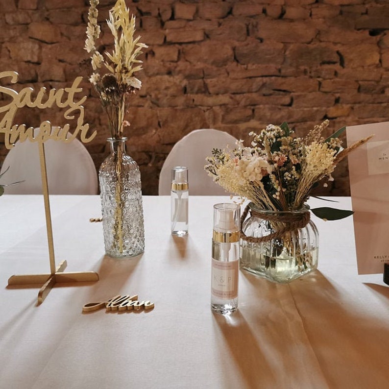 May include: A wedding table setting with a gold sign that says "Saint Amour" and a gold decorative stand. There are two clear glass bottles with dried flowers, two clear glass bottles with liquid, and a clear glass jar with dried flowers. The table is covered in a white tablecloth.