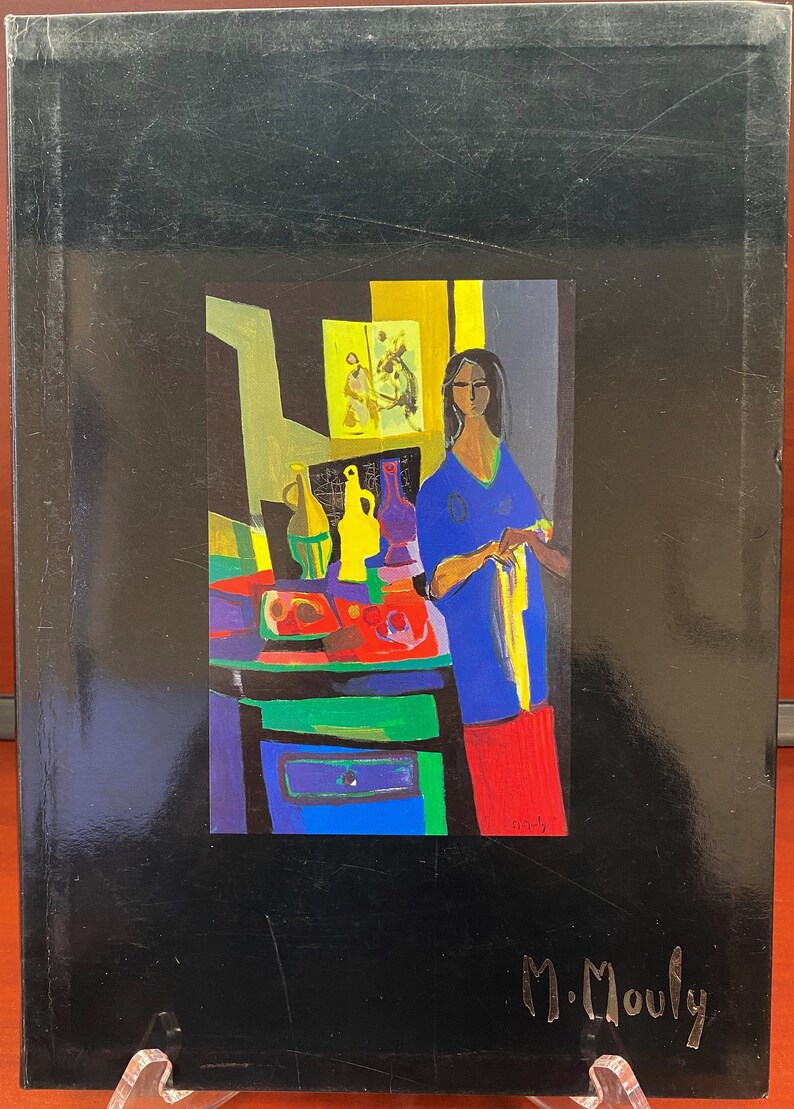 May include: A colorful painting of a woman standing in a room with a table and various objects. The woman is wearing a blue shirt and red pants. The painting is signed "M. Mouly".