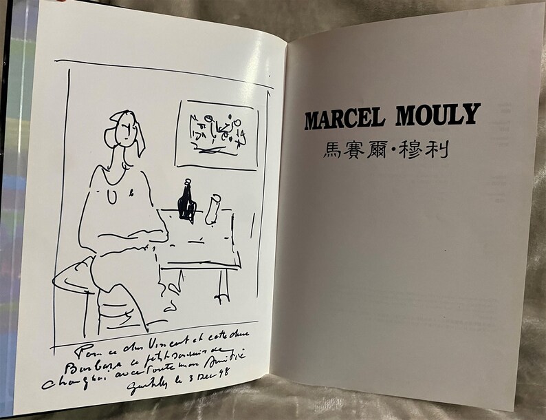 May include: A black and white sketch of a person sitting at a table with a bottle and a glass. The text "Pom u Am Vineant at catte chine Chaguri auce foute mon mits Zurhly le 3 dec 98" is written below the sketch. The text "MARCEL MOULY 馬賽爾&middot;穆利" is written in black on a white background.