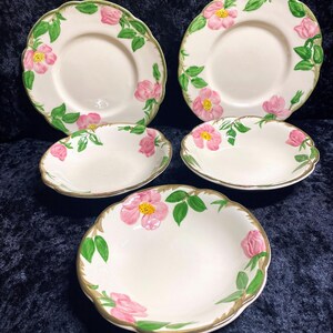May include: A set of vintage ceramic plates and bowls. The plates and bowls are white with a pink rose and green leaf design. The edges of the plates and bowls have a scalloped edge with a gold trim.