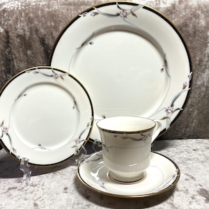 Può includere: Un set di stoviglie in porcellana bianca con bordo nero e dorato e un motivo floreale. Il set comprende un piatto piano, un piatto da insalata e una tazza da tè e un piattino.