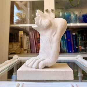 May include: A white sculpture of a human foot positioned vertically on a rectangular base. The sculpture displays detailed anatomical features including the toes, arch, and heel, rendered in a matte white finish with visible texture.