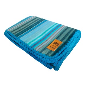 May include: A blue and green striped throw blanket with a crocheted border. The blanket has a small brown label with the text "The Alpaca People" on it.