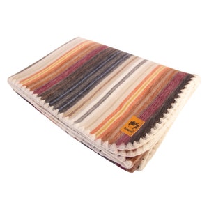 Puede incluir: Una manta de tiro a rayas con un borde de ganchillo en un esquema de colores crema, marrón, naranja y gris. La manta tiene una pequeña etiqueta que dice "Alpaca".