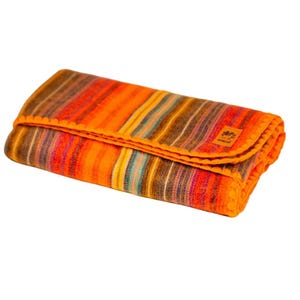 Puede incluir: Manta plegada con un vibrante estampado de rayas en tonos naranja, marrón, amarillo y rojo. La manta tiene un aspecto texturizado y un borde naranja decorativo. Una pequeña etiqueta es visible en la manta.