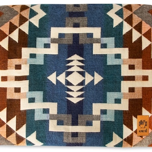 Puede incluir: Una manta de tejido azul, verde azulado, marrón y blanco con un patrón geométrico. La manta tiene un borde de flecos y una pequeña etiqueta con el texto "El Rito" en la esquina inferior derecha.