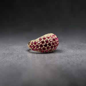 May include: A gold ring with a dome-shaped top, fully encrusted with numerous small, round, deep red gemstones. The ring's band is a polished gold color, and the gemstones are closely set together, creating a textured surface.