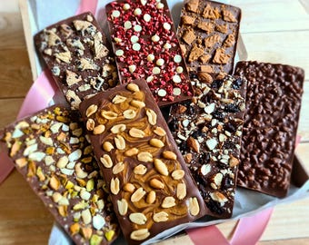 May include: Assortment of chocolate bars with various toppings, including peanuts, pistachios, dried fruit, and cookie pieces. The bars are arranged in a box with pink ribbon.