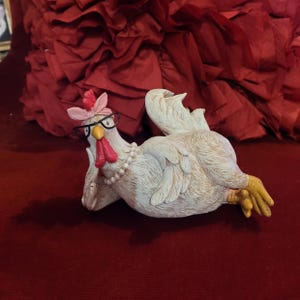 Puede incluir: Una figurita de pollo de cerámica blanca que lleva gafas rosas y una diadema rosa. El pollo está tumbado de lado con la cabeza apoyada en el ala. La figurita está sobre un fondo rojo.